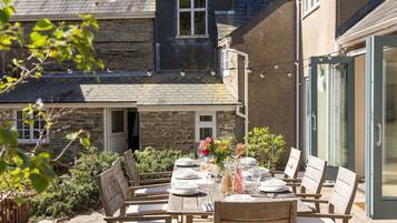 An outdoor dining area with a table set for six, surrounded by stone walls and a glass door leading to a garden.
