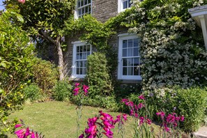 Ein Steinhaus mit weiß gefassten Fenstern, ein gepflegter Garten mit verschiedenen Pflanzen und Blumen sowie ein freier Blick auf die umliegende Grünfläche.