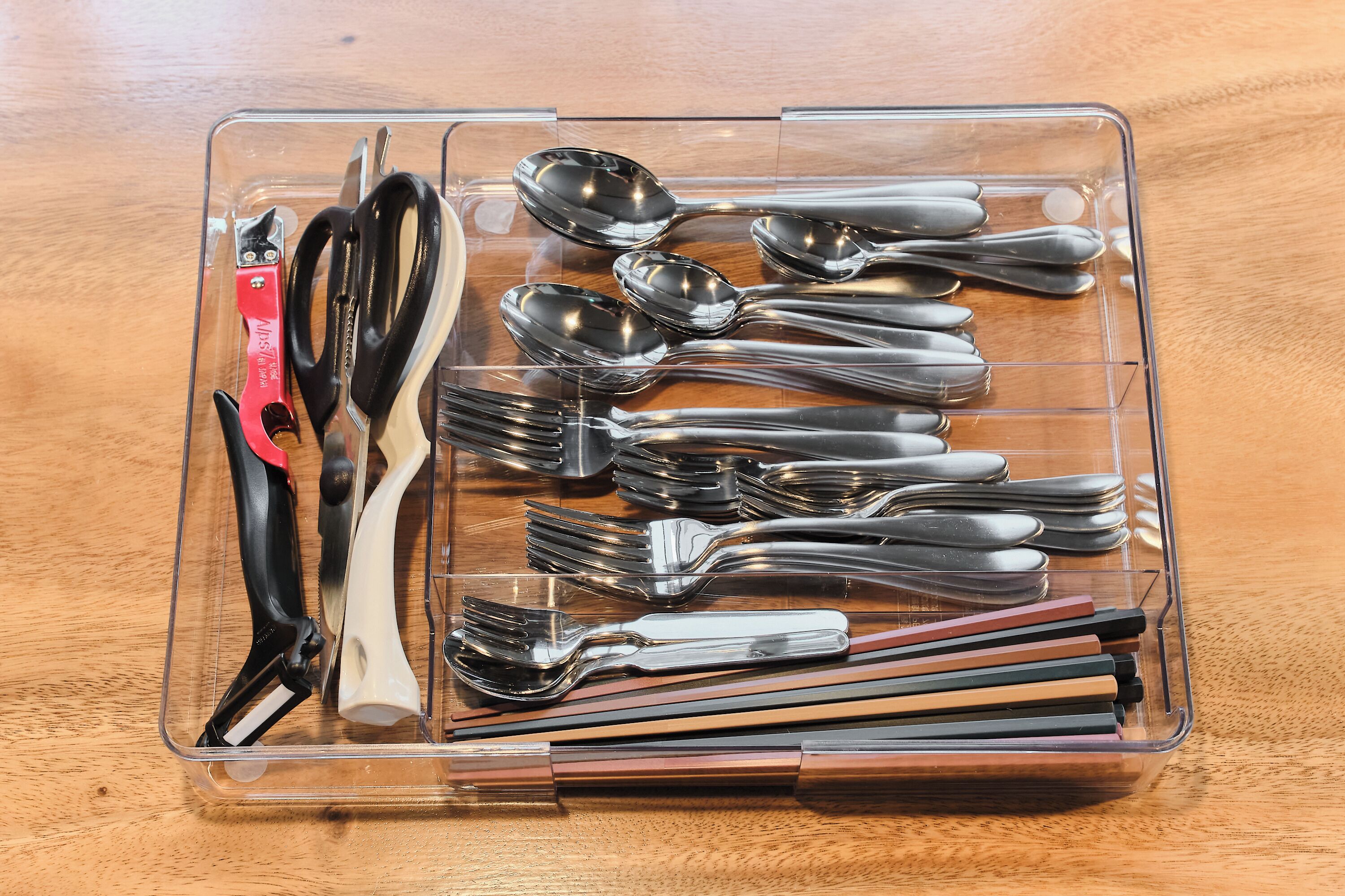A clear plastic storage box containing various utensils including forks, spoons, and a pair of scissors.