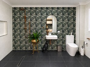 Een moderne badkamer met behang met een bladmotief, een gouden decoratieve plant, een witte wastafel, een toilet en een prullenbak.