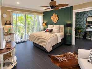 Een slaapkamer met een bed, een plafondventilator, een nachtkastje, een lamp, een dressoir, een raam met jaloezieën en uitzicht op groen buiten.
