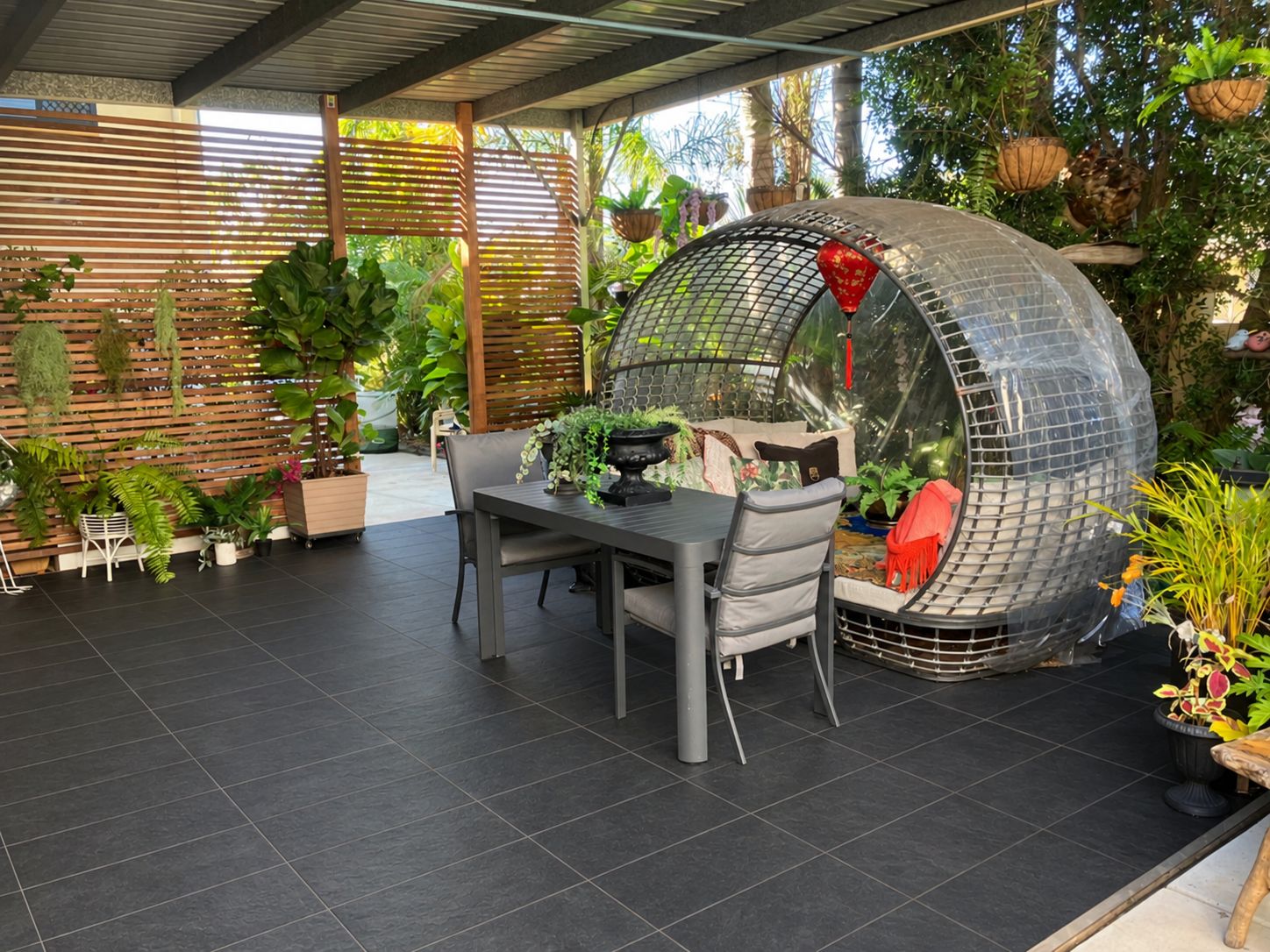 A covered patio area with a dining table and chairs, surrounded by potted plants and a unique spherical structure.