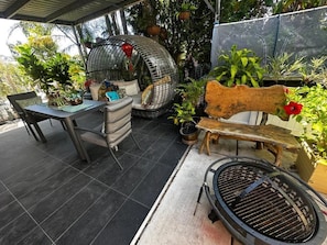 An outdoor patio with a grill, a wooden bench, a table with chairs, and various potted plants.