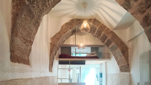 Eine gewölbte Decke mit einer Hängeleuchte, eine Couch mit Kissen und Blick in einen Raum mit Bett und Fenster.
