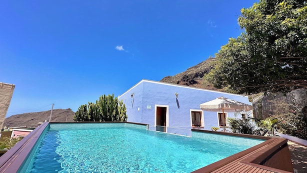 Un espace piscine avec un bâtiment bleu, une terrasse en bois et une chaîne de montagnes en arrière-plan.