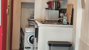 Een kleine keuken met een wasmachine, een afvalbakje en een koffiezetapparaat.