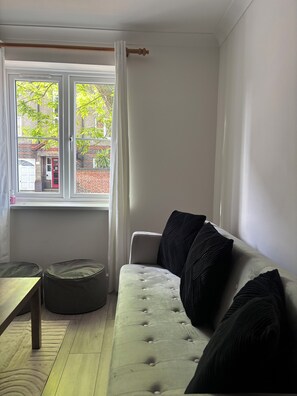 A living room with a sofa, cushions, a window with curtains, and a view of a building outside. - Modern London Stay for 5|Parking| Excel, City Airport & Canary Wharf (London)