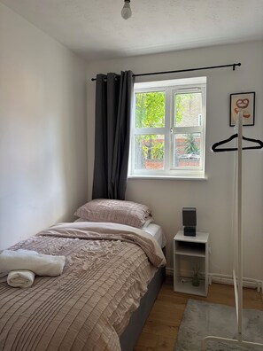 A bedroom with a bed, a window, a radiator, a small table with a speaker, and a wall-mounted coat rack. - Modern London Stay for 5|Parking| Excel, City Airport & Canary Wharf (London)