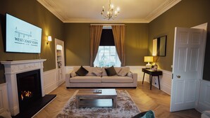 A living room with a fireplace, a sofa set, a coffee table, and a television displaying 'WEST COAST STAYS LTD.' - Bankhouse Troon, Scotland | Luxury Sea View Retreat with Hot Tub & Sauna (Troon)
