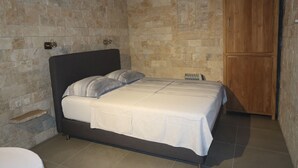 Un dormitorio con pared de piedra, cama con ropa blanca, armario de madera y una pequeña estantería.
