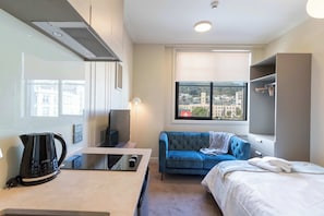 A modern hotel room with a kitchenette, a blue sofa, a bed, and a large window with a city view. - Te Aro City Stay - Modern Studio for Two (Wellington)