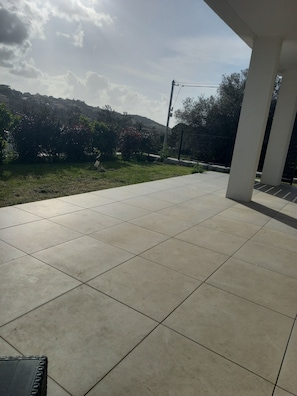 Un patio couvert doté d’un sol carrelé, offrant une vue sur un jardin aménagé et sur une colline située au loin.
