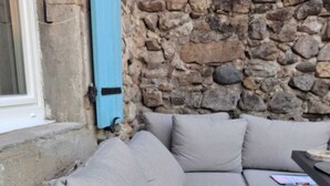 Rincón con asientos, pared de piedra, ventana con persianas azules y una mesa con un libro.