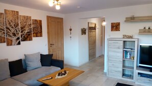 Uma sala de estar com um sofá, uma mesa de centro com pinhas, um rack para televisão com livros e uma porta de madeira.