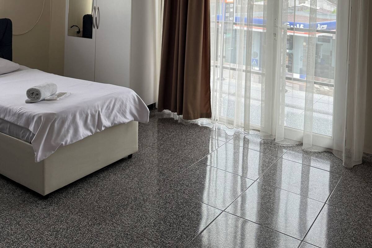 A hotel room with a bed, a wardrobe, a mirror, an air conditioning unit, and a view of the outside through a large window with curtains.