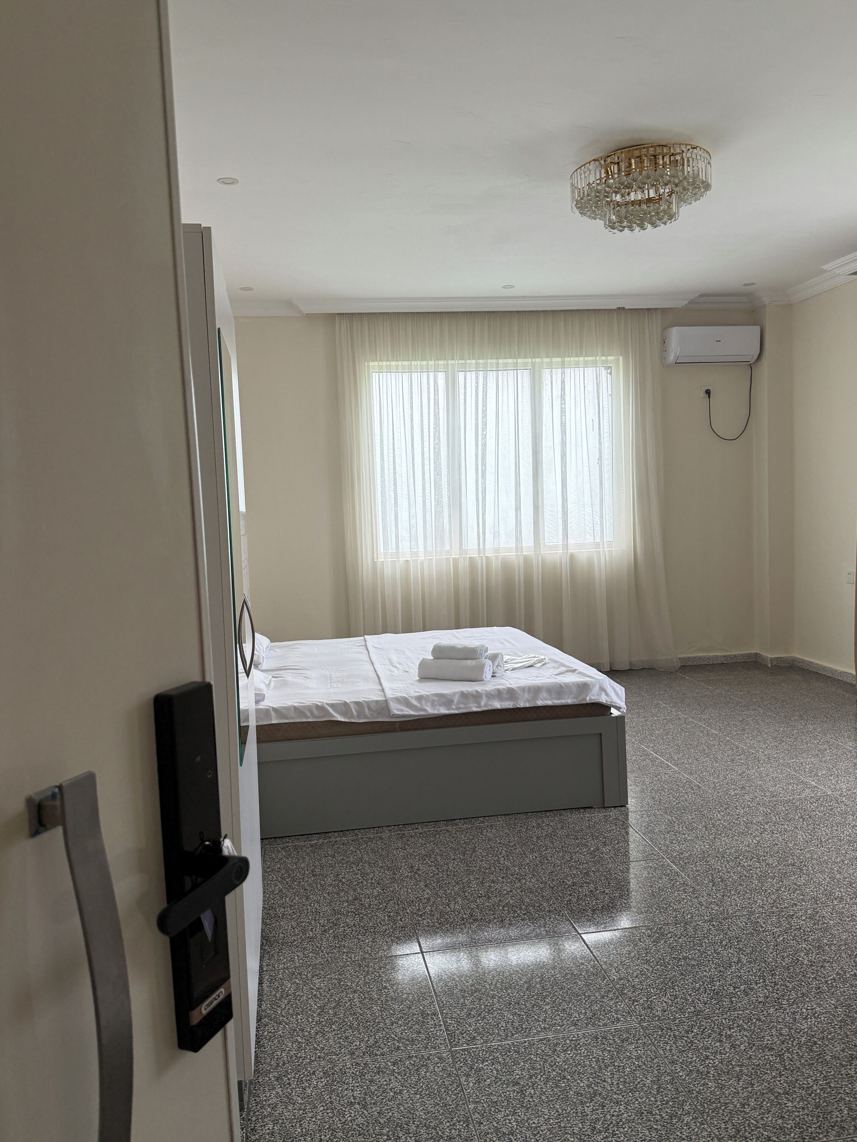 A hotel room with a bed, a ceiling-mounted air conditioner, and a large window with curtains.