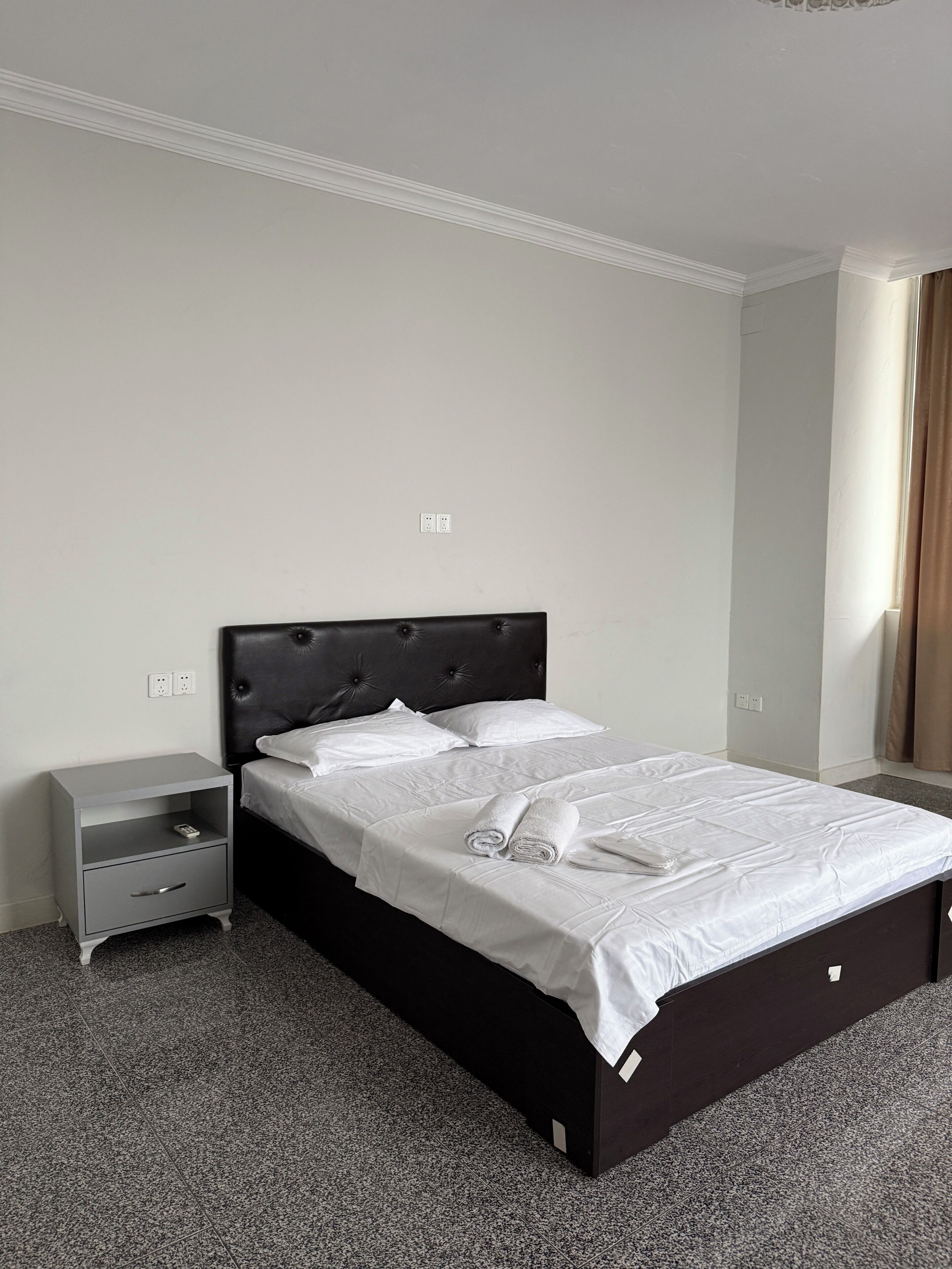 Ein ordentlich bezogenes Bett in einem Hotelzimmer mit dunklem Kopfteil, einem kleinen Nachttisch und einer Steckdose an der Wand.