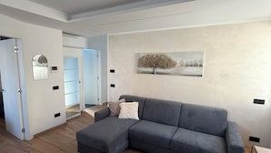 En moderne stue med en grå hjørnesofa, et lille bord og en stol. Et billede hænger på væggen, og en dør fører til et andet rum.