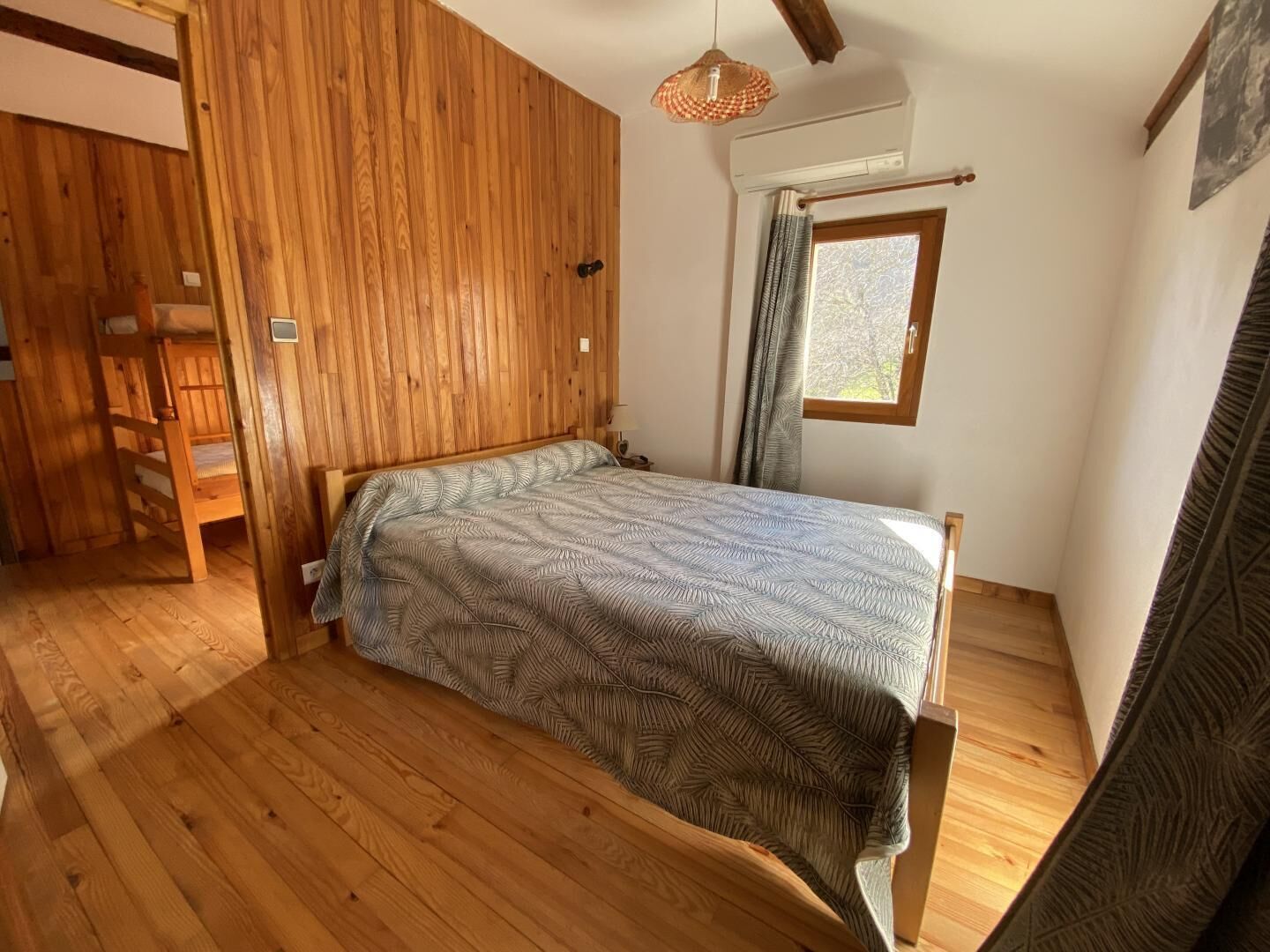 Une chambre avec un plancher en bois, un lit recouvert d’une couvre-lit à motifs, une fenêtre avec des rideaux et un luminaire suspendu.