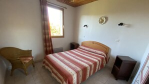 Une chambre à coucher comprenant un lit, une chaise, une petite table, une fenêtre avec des rideaux et un luminaire fixé au mur.