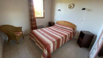 Une chambre à coucher comprenant un lit, une chaise, une petite table, une fenêtre avec des rideaux et un luminaire fixé au mur.