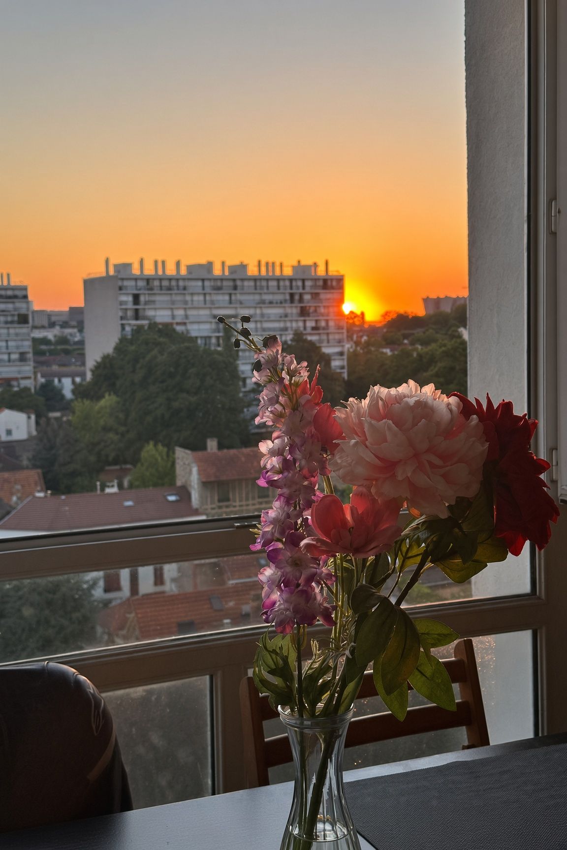Een vaas met bloemen op een tafel, bij een raam met uitzicht op een stadslandschap in de zonsondergang.