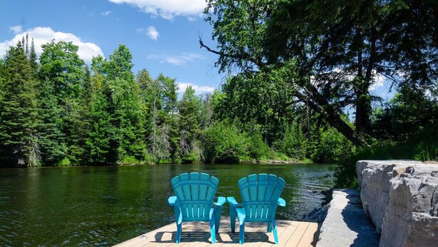 A wooden dock with two chairs facing a calm lake surrounded by dense forest. - Rustic River Front Cottage Cozy*Kayaks*Hot Tub*Games (Kawartha Lakes)