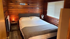 A wooden cabin room with a bed, a nightstand with a lamp, a fan, and a window with blinds. - Rustic River Front Cottage Cozy*Kayaks*Hot Tub*Games (Kawartha Lakes)