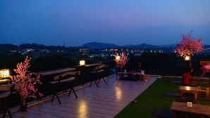 An outdoor terrace with wooden furniture, a view of distant city lights, and cherry blossom trees. - Sakura Yama Resort (Khaem Son)