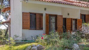 A single-story building with a tiled roof, wooden shutters, and a garden with rocks and plants. - Villa Anty Holiday Home in Sani, Chalkidiki—Private Terrace & Pine Forest Views (Sane)