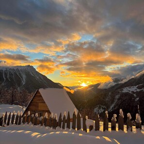 Eine Holzhütte mit geneigtem Dach, umgeben von einem verschneiten Zaun, vor einer Berglandschaft bei Sonnenuntergang.