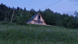 Une maison en bois d’un seul étage, avec un toit en pente, entourée d’une clôture en bois, située dans un champ herbeux avec des arbres en arrière-plan.
