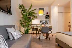A modern living room with a sofa, a dining table, chairs, a large plant, and a kitchen area in the background.