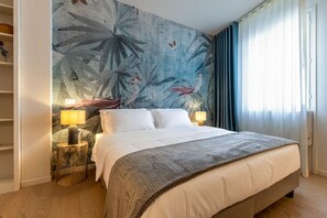 Hotellihuone, jossa on suuri sänky, kaksi yöpöytävalaisinta, seinämaalaus ja ikkuna, jossa on verhot.
