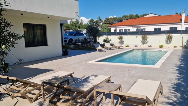 Una piscina con sillas de descanso, un edificio blanco y una casa con techo rojo al fondo.