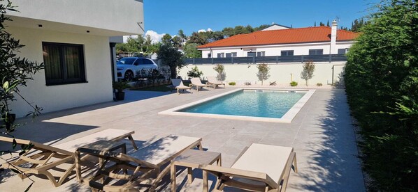 Ein Schwimmbecken mit Liegestühlen, ein weißes Gebäude und ein Haus mit rotem Dach im Hintergrund.