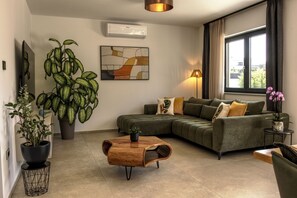 Ein modernes Wohnzimmer mit Ecksofa, Couchtisch, großer Topfpflanze, Wandklimaanlage und einem Vorhangfenster.