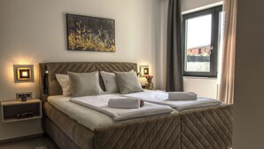 Una cama doble con cabecera acolchada, mesitas de noche con lámparas, un cuadro en la pared y una ventana con cortinas.