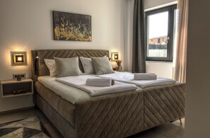 Ein Doppelbett mit gestepptem Kopfstück, Nachttische mit Lampen, ein gerahmtes Bild an der Wand und ein Fenster mit Vorhängen.