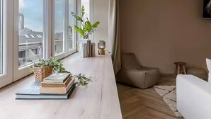 A modern living room with large windows, a long wooden table, a white sofa, and a view of the cityscape. - Juan | Trendy Renovated Apartment in the Heart of Ostend | Sleeps 8 (Oostende)