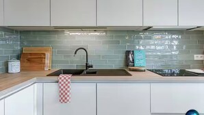 A modern kitchen with white cabinets, a black faucet, a wooden cutting board, and a checkered towel. - Juan | Trendy Renovated Apartment in the Heart of Ostend | Sleeps 8 (Oostende)