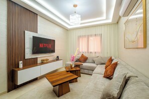 Ein modernes Wohnzimmer mit einem Flachbildfernseher, Holzmöbeln, einer Deckenleuchte und einem großen Fenster mit Vorhängen.