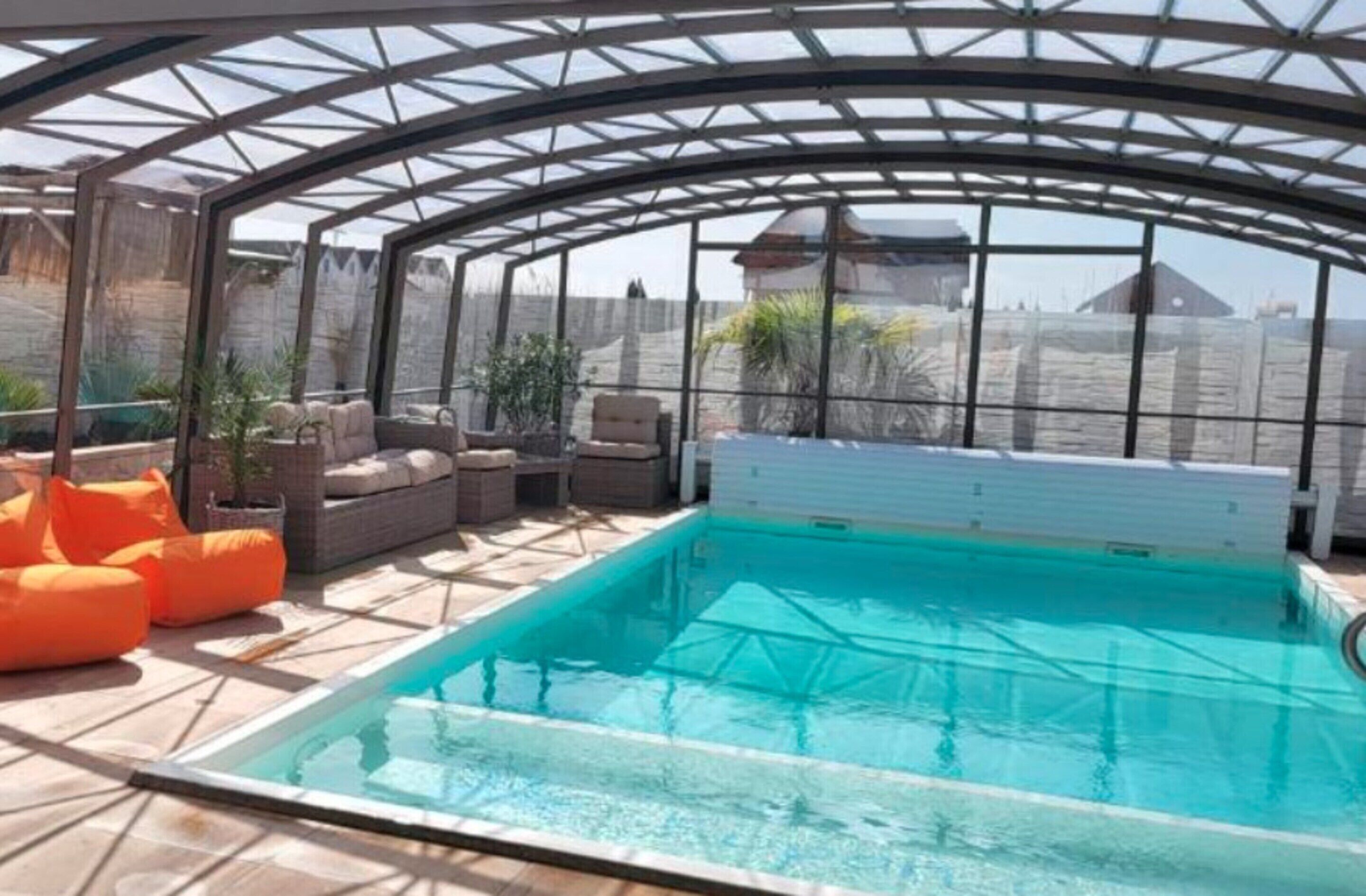 A covered swimming pool with a glass ceiling, surrounded by seating and potted plants.