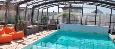 A covered swimming pool with a glass ceiling, surrounded by seating and potted plants.