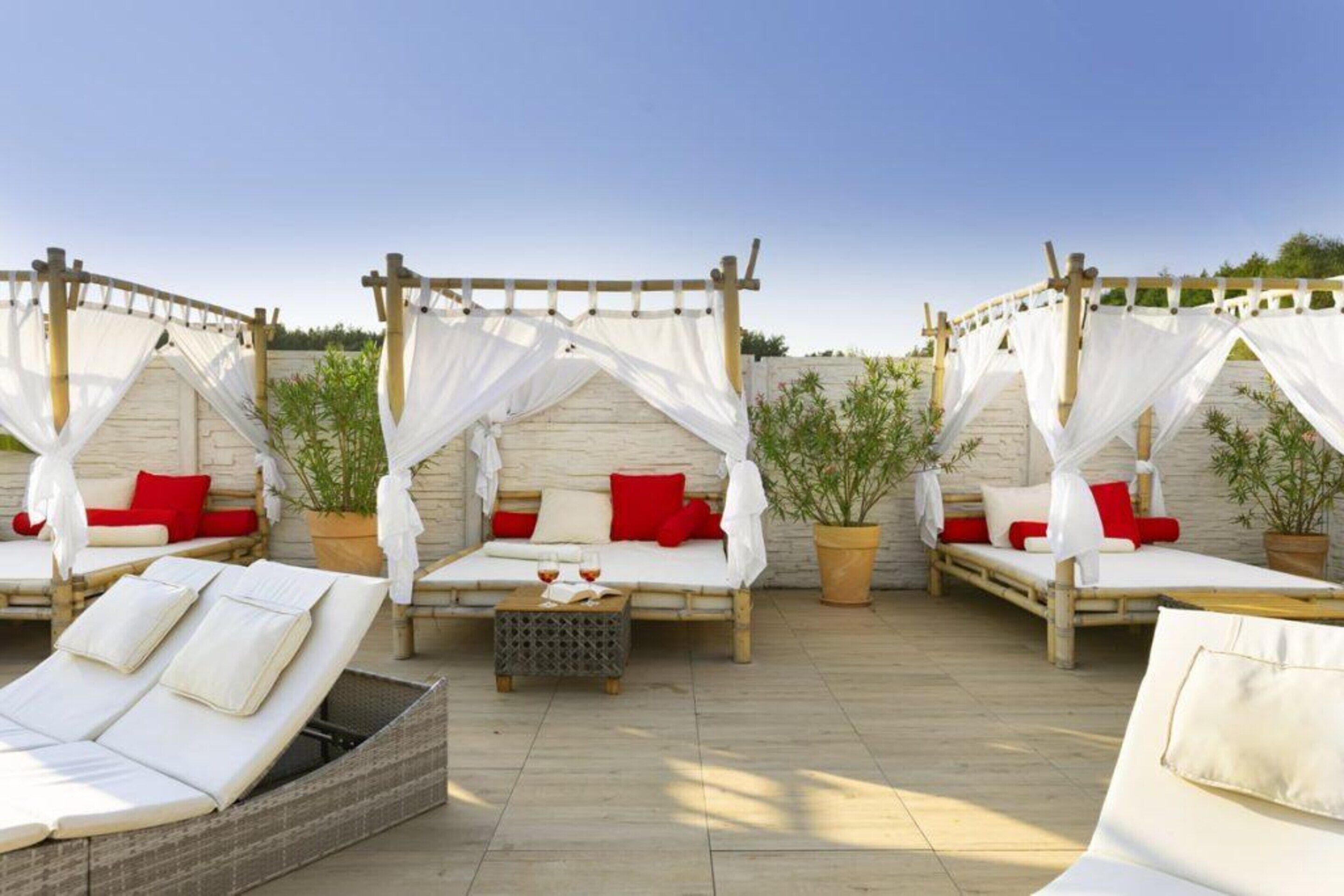 A rooftop lounge with four wooden beds, each with a canopy and red pillows, surrounded by potted plants.