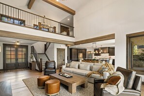 A spacious living area with a high ceiling, a staircase to an upper level, a large sofa set, and a kitchen in the background. - Whisper Ridge - Luxury 5 BD - Walk to Social Village (CLE ELUM)