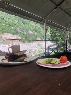 Une table dressée avec de la nourriture et une tasse, donnant sur une rivière et des collines verdoyantes.
