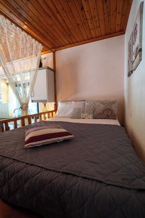 A bedroom with a wooden ceiling, a bed with a dark quilt, and a window with a lace curtain.