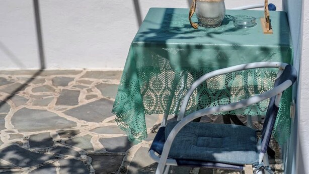 Une table recouverte d’une nappe verte, une carafe en verre et une chaise posées sur un sol en pierre, avec vue sur la mer.