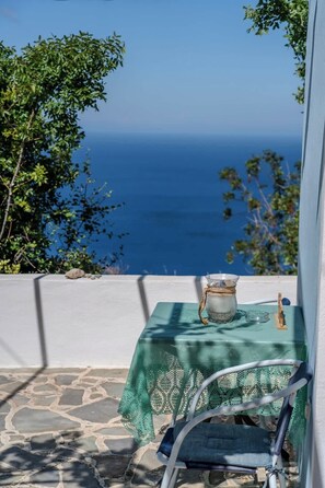 A table with a green tablecloth, a glass pitcher, and a chair on a stone floor with a view of the sea.
